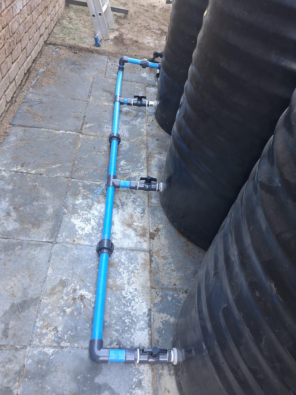 Row of black water tanks linked by blue manifold pipework and ball valves